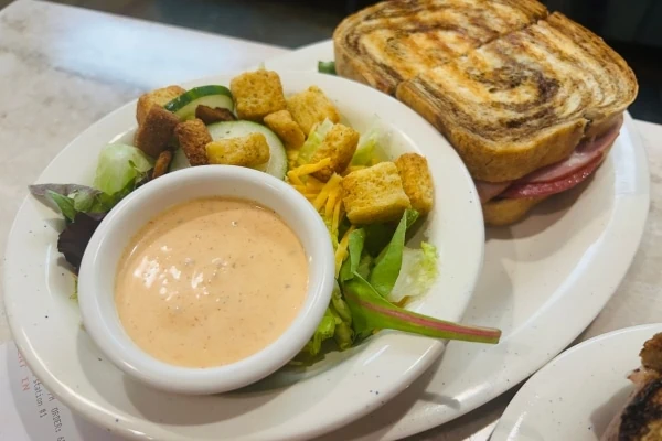 A lunch sandwich served with a salad and dressing, on a plate.