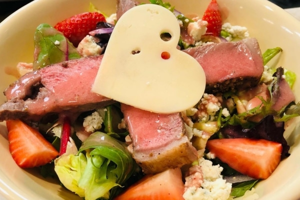 A salad with steak and strawberries served with a heart shaped piece of cheese on top.